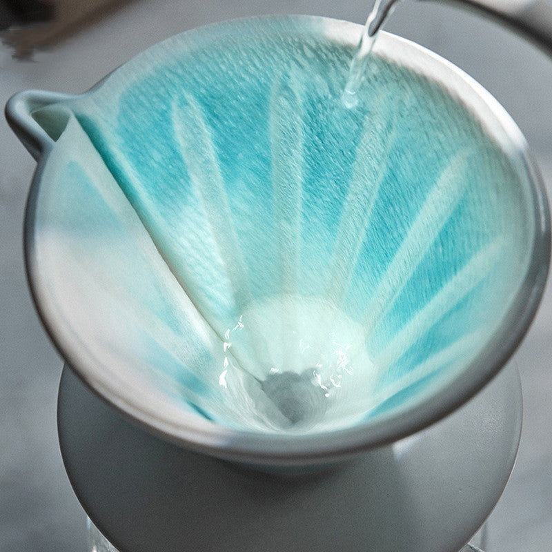 Tea filter with blue liquid being poured into a white teapot on a gray surface