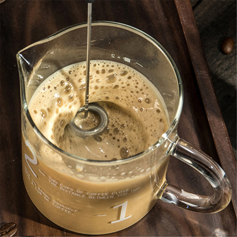Glass measuring cup with coffee and a frother on a wooden surface