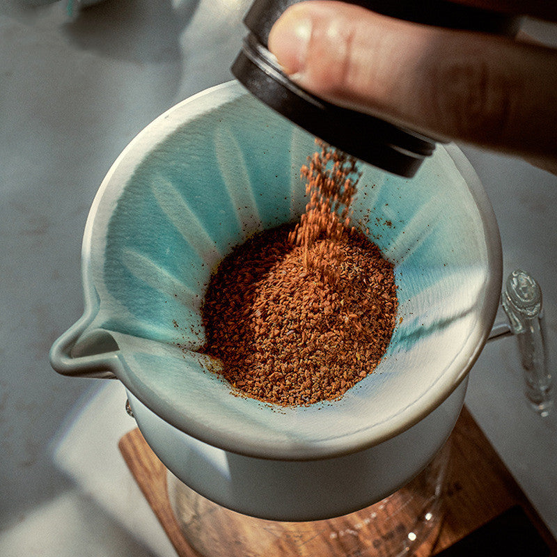 Hand pouring coffee grounds into a white ceramic filter with a blue rim.