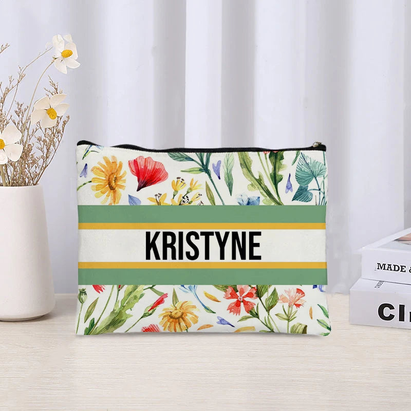 Floral-patterned pouch with 'KRISTYNE' printed on it, placed on a surface with a white curtain and potted plant in the background.
