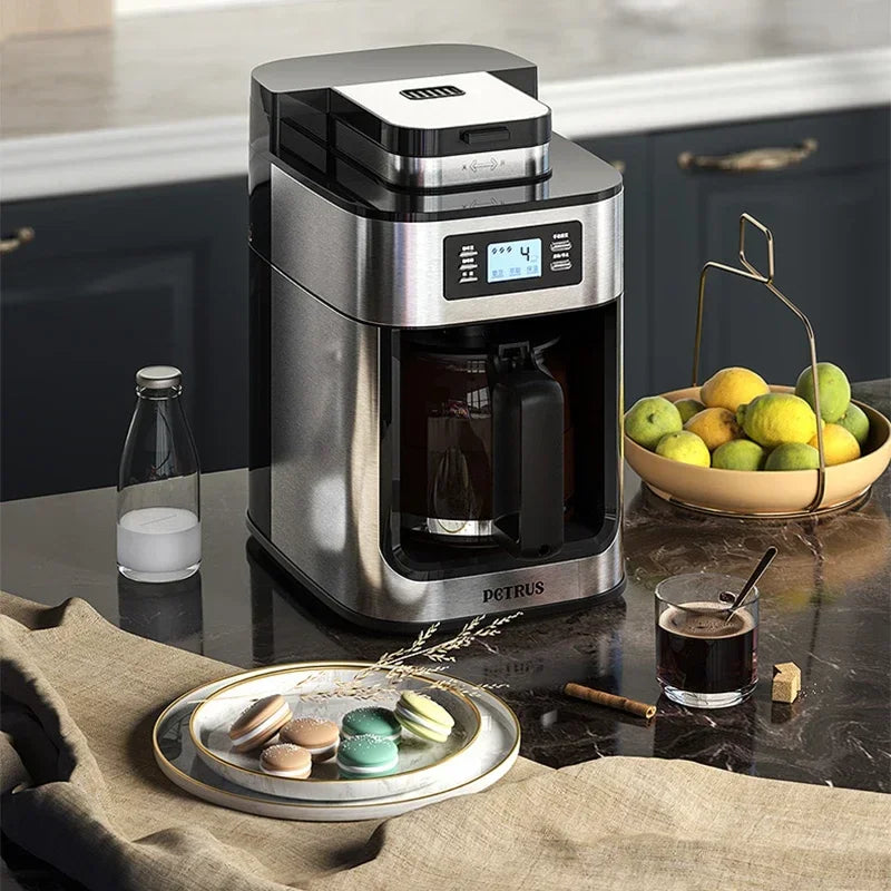 Coffee maker on a kitchen counter with a glass of coffee and fruit bowl in the background