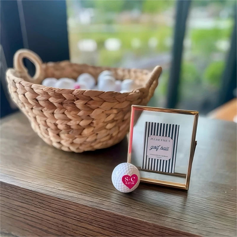 Personalised golf balls UK - A small white golf ball with a pink heart logo sits in front of a gold-framed sign that reads 'please see a golf ball,' with a woven basket of golf balls in the background on a wooden table.