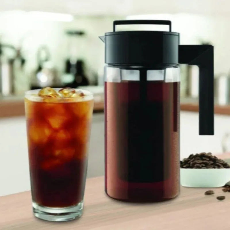 Glass carafe filled with iced coffee next to a glass of iced coffee on a wooden surface.