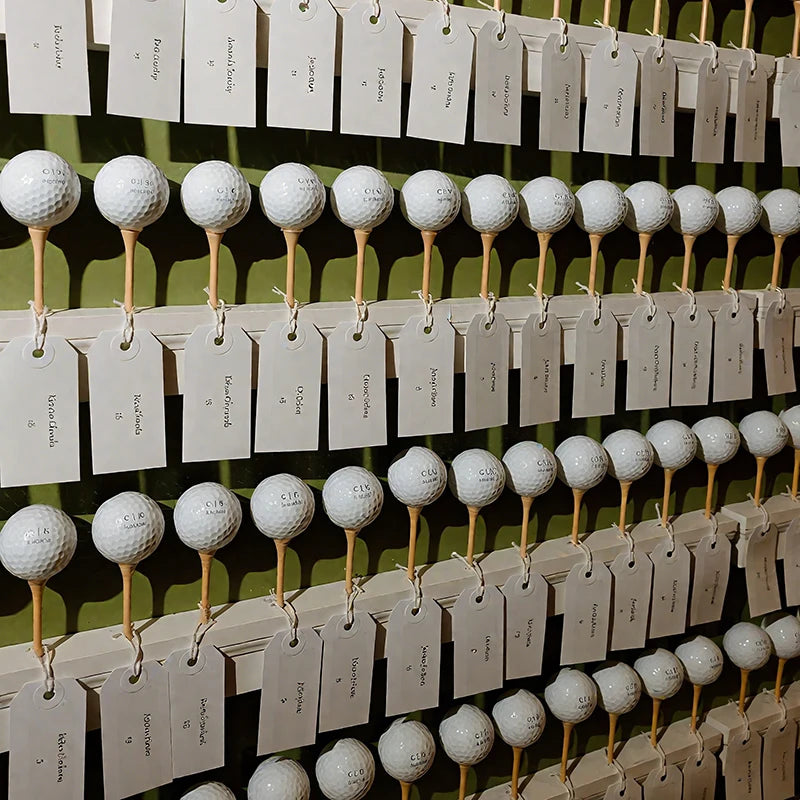 Personalised golf balls UK - Rows of white golf balls on wooden sticks, each attached to a small paper tag, displayed against a dark olive-green wall.