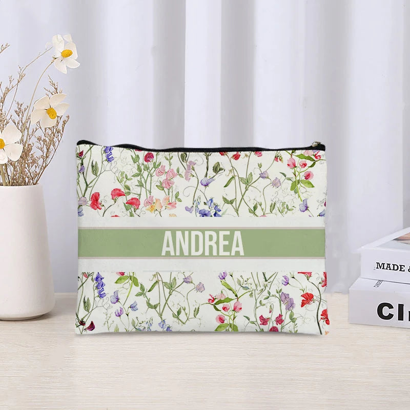 Floral-patterned pouch with 'ANDREA' printed on it, placed on a surface with a white curtain and flowers in the background.